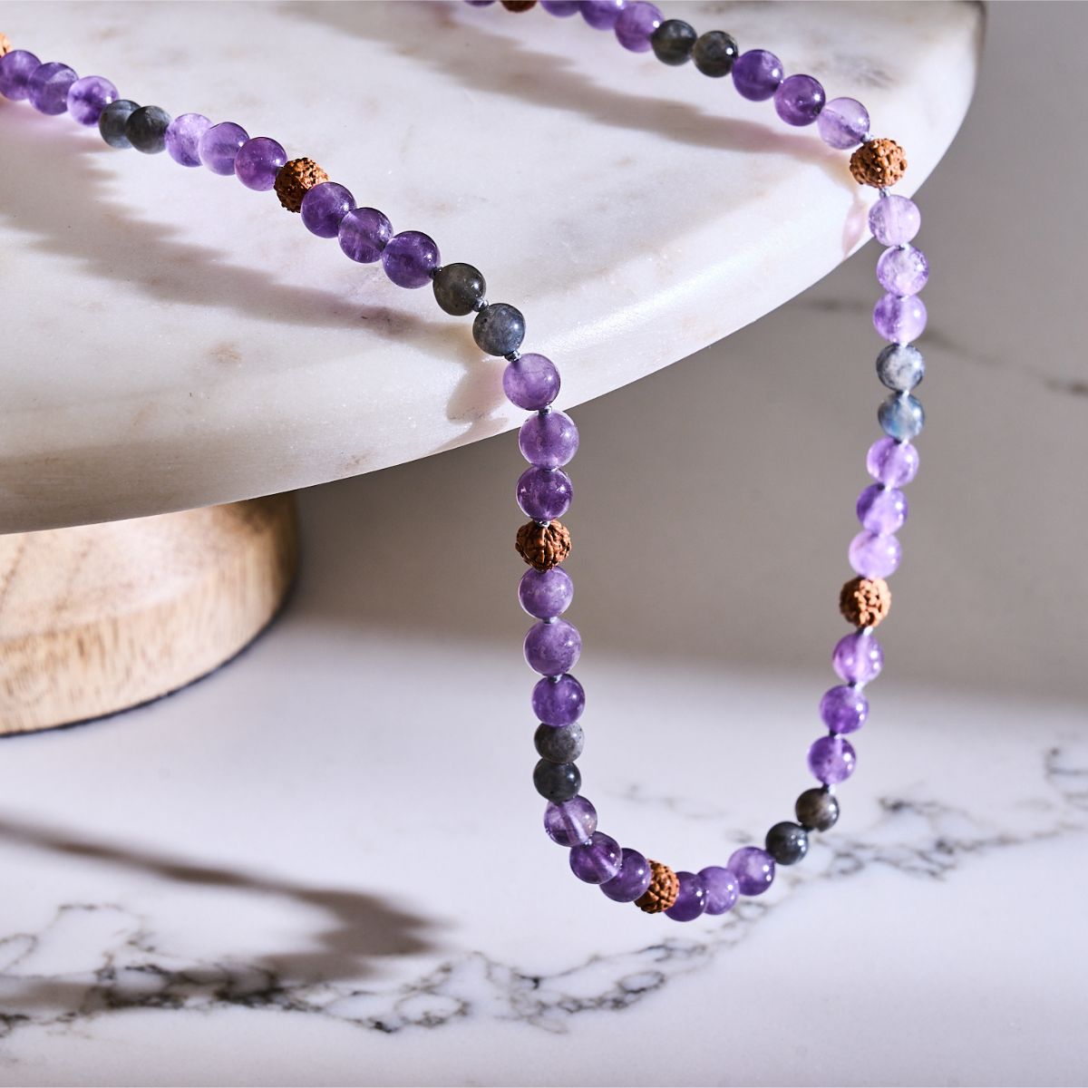 Purple amethyst and dark labradorite beaded necklace with textured brown seed beads, draped over a marble pedestal.