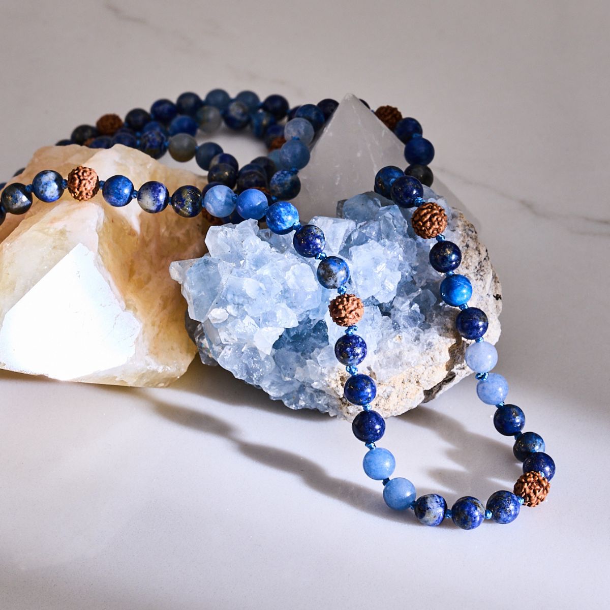 Blue beaded gemstone mala necklace with natural brown rudraksha seed accents, draped over pale crystal clusters.