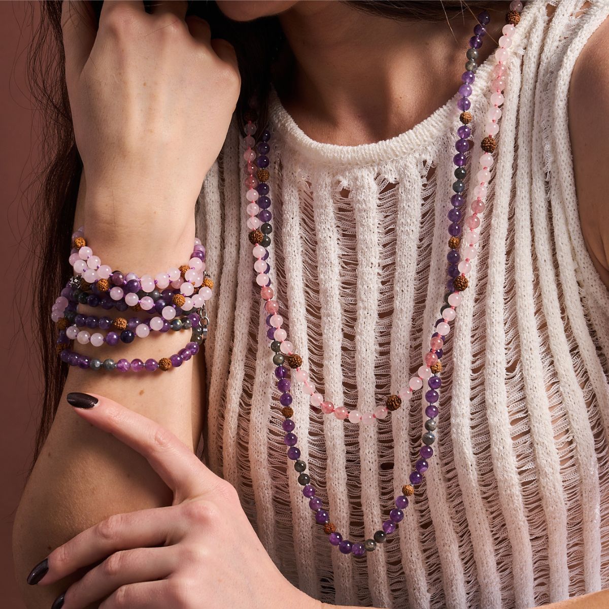 Layered purple, pink and gray beaded bracelets and long necklaces styled with a white open-knit top.