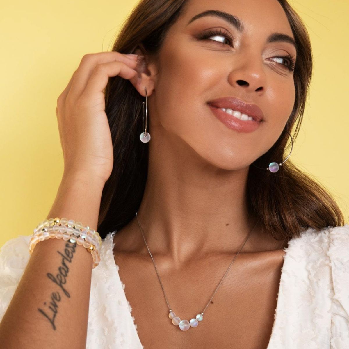 Model wears a delicate silver-tone necklace and long drop earrings with iridescent pearl-like beads, paired with beaded bracelets.