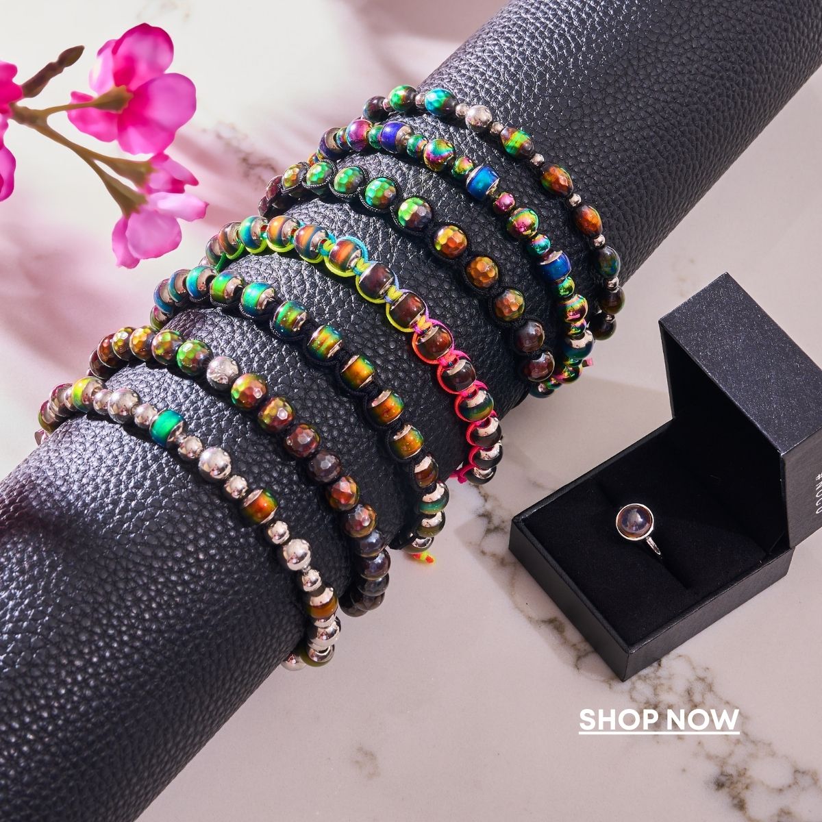 NOGU multicolor iridescent beaded bracelets wrap a black display, styled with pink flowers and a boxed gemstone ring.