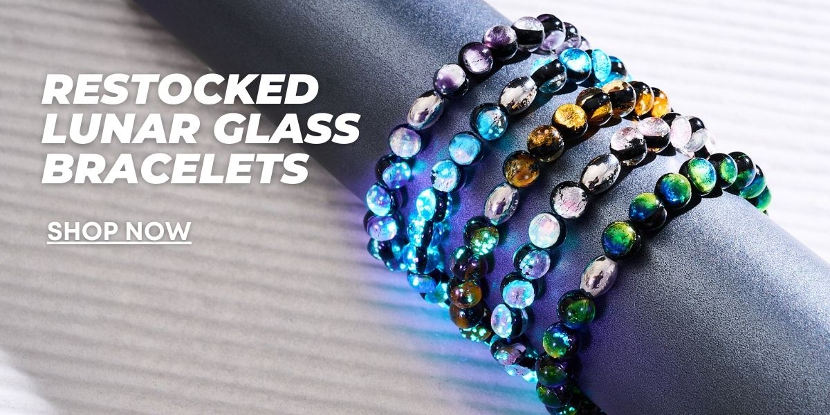 Stacked Lunar Glass beaded bracelets in iridescent blue, green, purple, gold, and silver shimmer on a wrist.