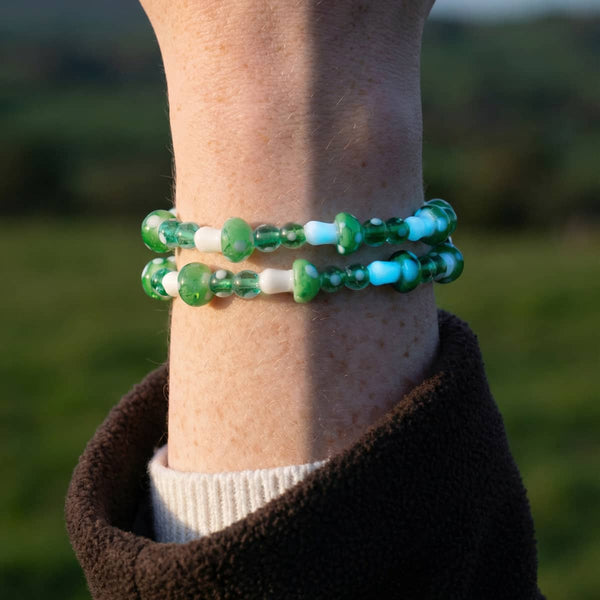 Green and white glow-in-the-dark Firefly glass mushroom bracelet on clear stretch cord, shown layered on a wrist.
