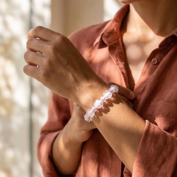 A close-up lifestyle shot of a person wearing a translucent pink beaded bracelet on their wrist, wearing a rust-colored linen shirt.