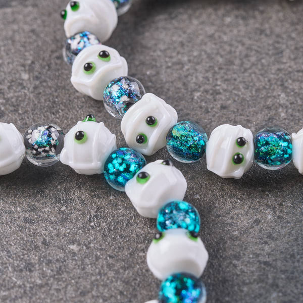 Teal Crypt Firework Glass Mummy Bracelet with white mummy beads, green eyes, and shimmering teal blue glass accents.